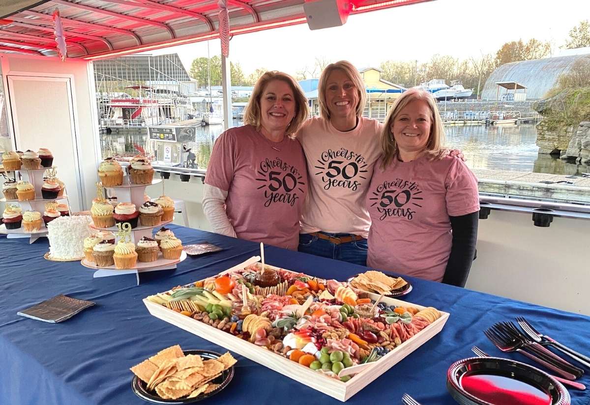 three ladies in matching 50th birthday shirts standing behind food spread